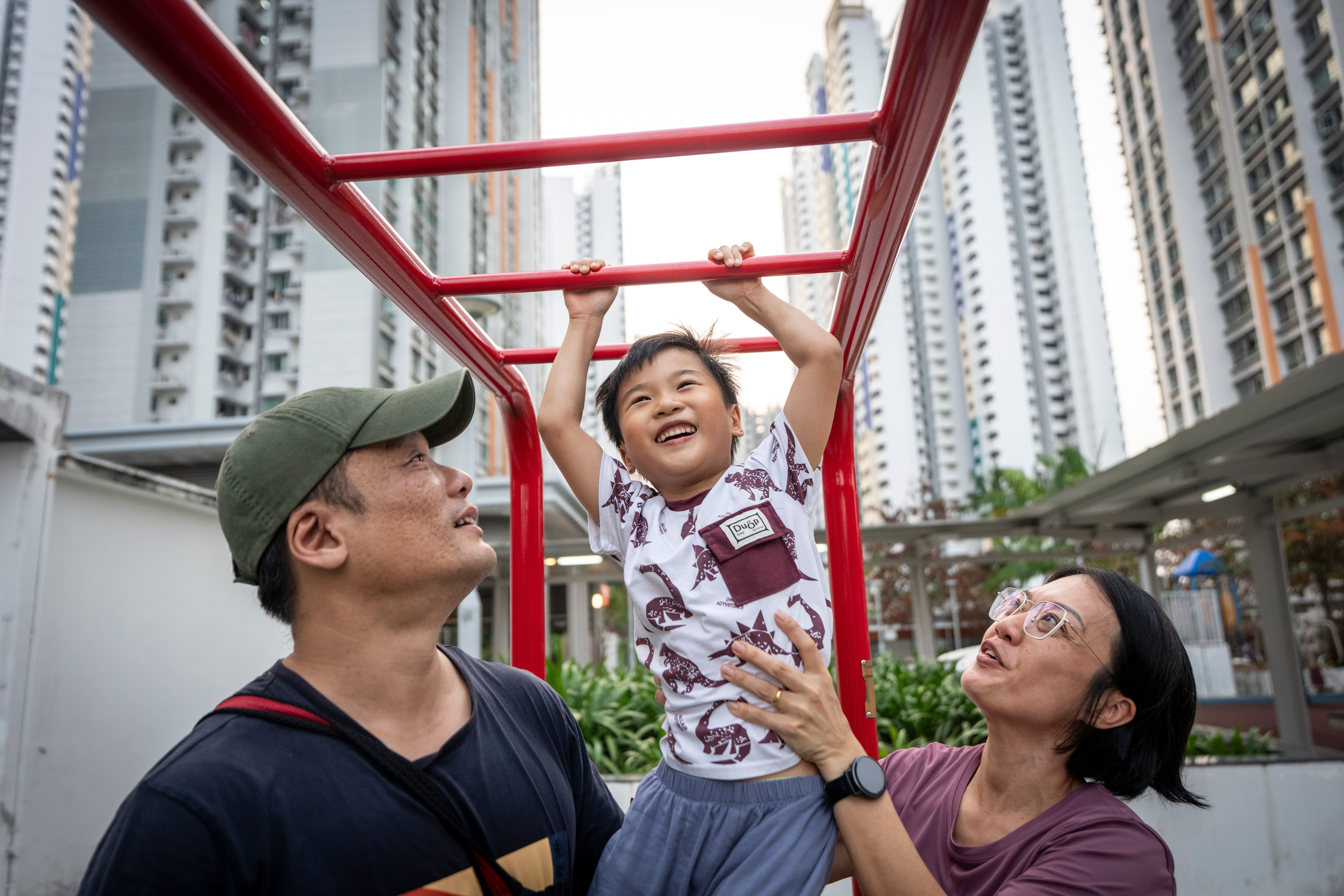Parents lifting their young son up to the monkey bars at a playground, all three smiling.